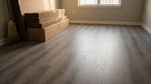 A vacant apartment living room with a new grey vinyl plank floor and flooring boxes stacked by the wall